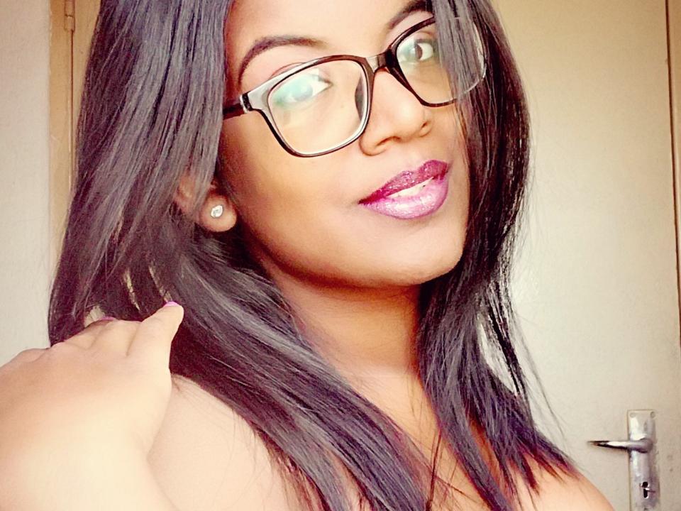 IndianBeaut with dark straight hair, glasses, shiny purple lipstick, and earrings, touching her hair, smiling. Her hand is near her head. She's wearing a strapless top.
