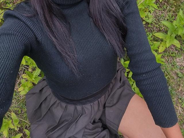 KiriamGomez has dark, ribbed, long-sleeve top, showing small, perky tits, and is wearing a black skirt, with long, black hair, and tan legs, in a grassy area.