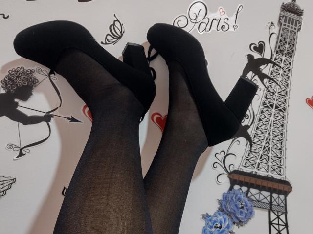 As pernas de Kiriam Gomez, vestindo meias finas pretas e sapatos de salto alto pretos com tiras nos tornozelos, com os pés cruzados, em frente a uma parede com tema parisiense..