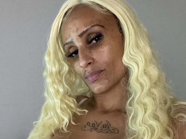 SexyRedBone with long, wavy blonde hair, dark skin, freckles, purple eyeliner, and a "Vali" tattoo on her chest, looking at the camera with a subtle, confident smile.