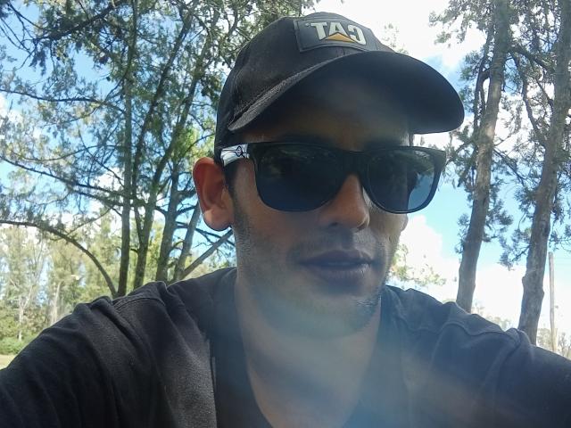 Alendro, wearing a black TA cap and dark sunglasses, with a beard and black shirt, in a sunlit forest. His ass is not visible. His face is in focus.