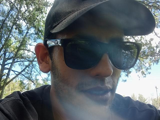Alendro with dark sunglasses, black cap, and black shirt, showing a slight mustache and stubble, standing outdoors with sunlight reflecting off his glasses.