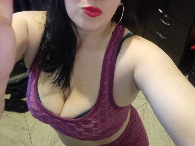 CharloteEzra tiene senos grandes, lleva un sujetador deportivo morado con estampado y un sujetador verde debajo, lápiz labial rojo y cabello negro, y está de pie en una habitación con pisos de madera..