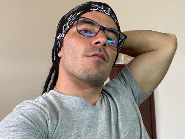 VictorPi, wearing a black and white bandana, gray tee, and glasses, with a relaxed, slightly smug expression, has a hand behind his head, showing his ass.