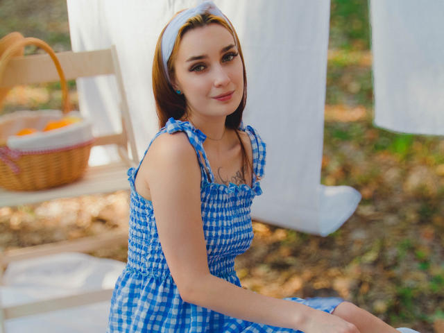 AlexisHott with brown hair, wearing a blue and white checkered dress, white headband, small tits, sitting outdoors, smiling, basket with oranges and white cloth in background.