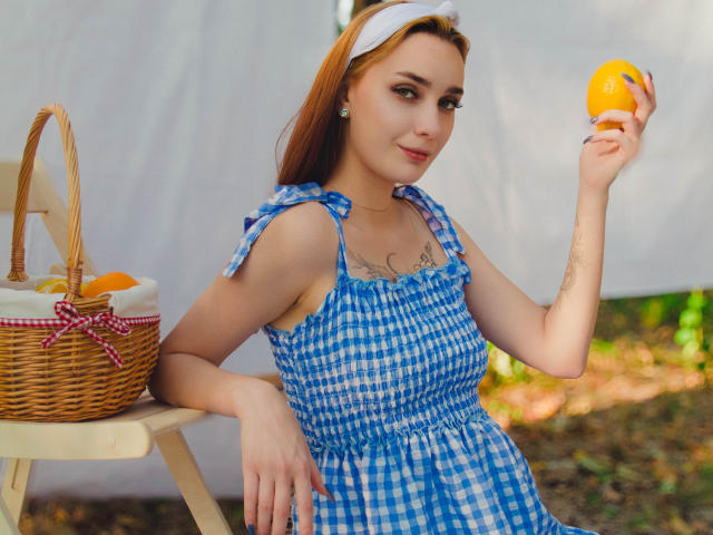 AlexisHott with red hair, white headband, blue check dress, holding orange, basket with apples and oranges on wooden table, tattoo on chest, sitting, outdoor, daytime.
