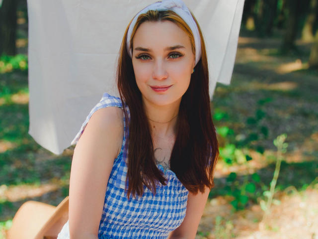 AlexisHott with brown hair, blue checkered dress, white headband, smiling at camera, tits visible, sitting outdoors, green foliage in background, sunlight on face, cute, sexy, relaxed.