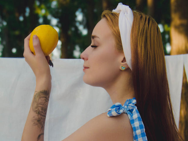 AlexisHott with red hair, white headband, blue check halter, holding yellow lemon, tattoo on right arm, earrings, eyes closed, side profile, holding lemon near nose.