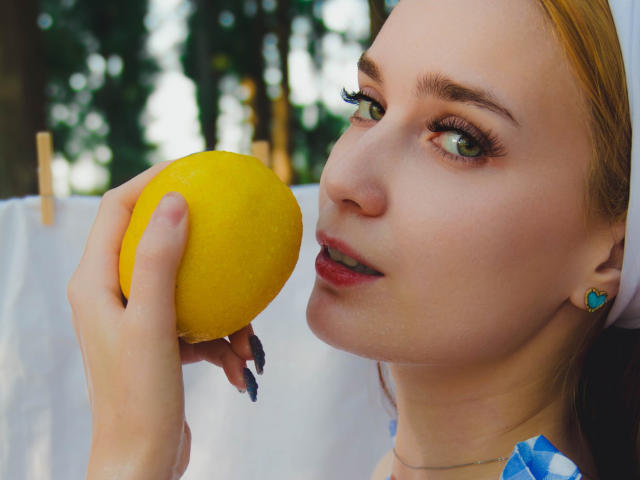 AlexisHott with green eyes, holding a yellow lemon to her mouth, wearing blue earrings, has light skin, and is looking slightly to the side with a subtle smile.