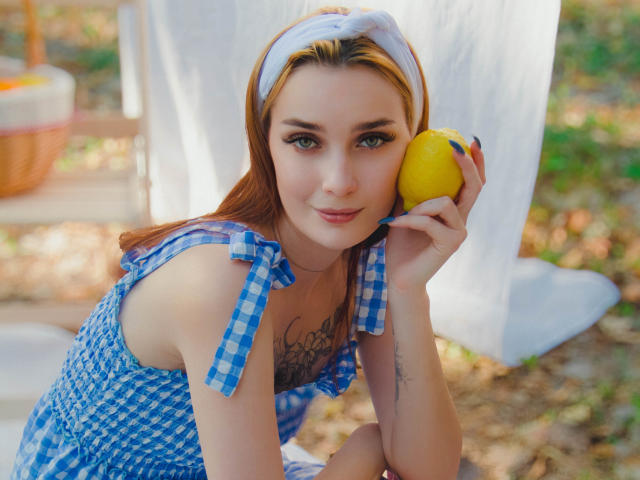 AlexisHott with red hair, blue check dress, white headband, holding yellow lemon, tattoo on chest, sitting outdoors, looking at camera, smiling slightly, bright sunlight, cute, sexy.