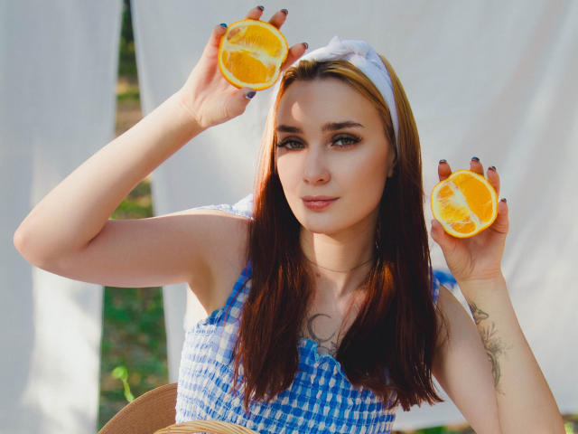 AlexisHott with long brown hair, blue checkered dress, holding two orange halves above her head, white headband, tattoo on chest, looking at camera, sitting outdoors, nails painted black.