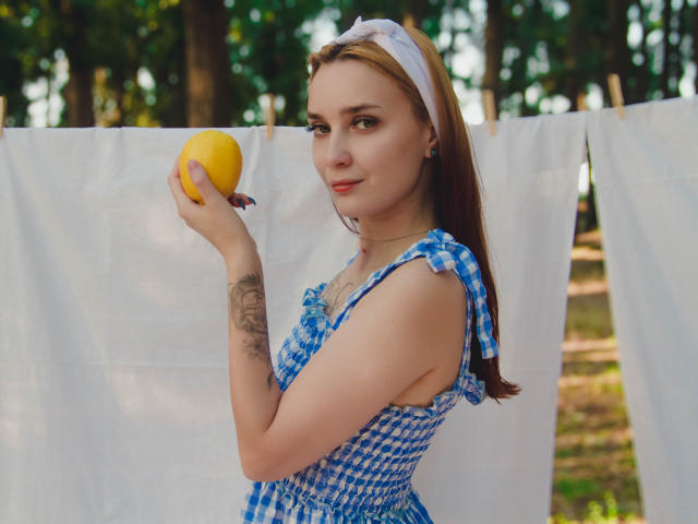 AlexisHott holding a yellow lemon, wearing a blue checkered dress with ruffles, white headband, and a tattoo on her right arm, standing in front of a white laundry line.