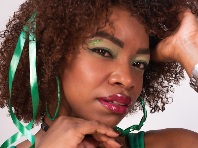DannaBanks, con cabello castaño rizado, maquillaje de ojos verde, labios rosados brillantes, un lazo verde, aros grandes y una mano en su barbilla, mirando ligeramente hacia un lado..