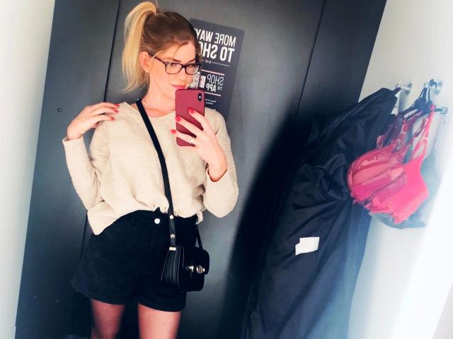 Tara-Secret-hot with blonde ponytail, glasses, white sweater, black shorts, black purse, holding pink phone, standing in front of mirror, pink bra on hanger to right.