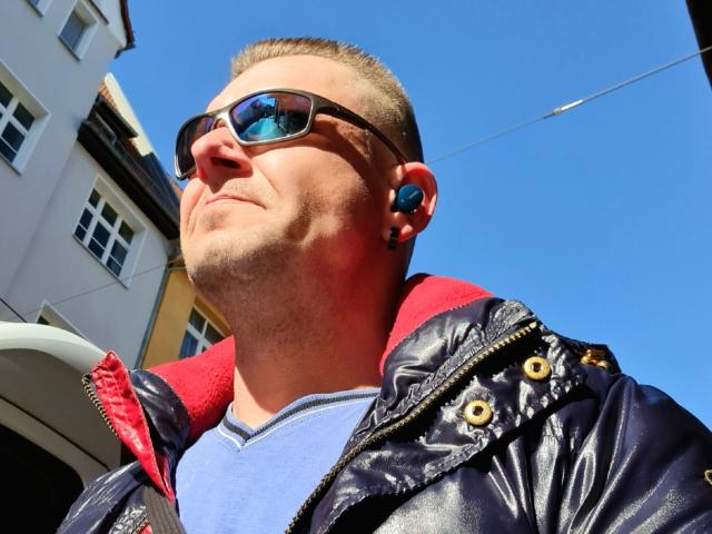 FaberShumi has a puffy dark blue jacket with yellow buttons, blue earbuds, sunglasses, and a short buzzcut. He's wearing a blue shirt and looking up. Clear blue sky.
