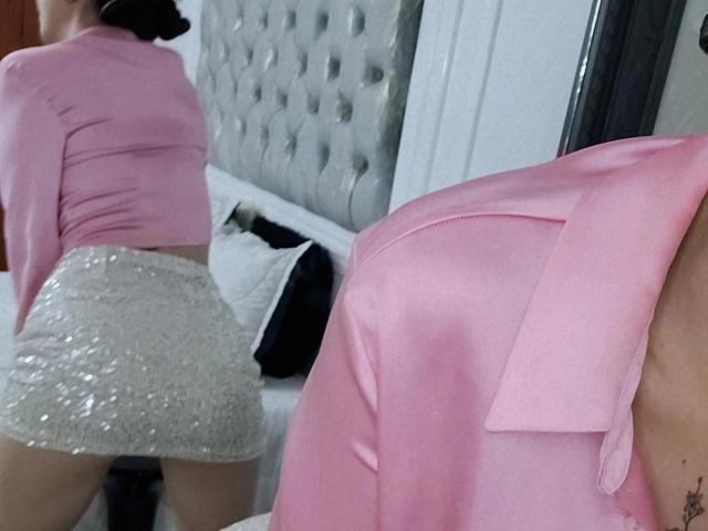 Emma Wells con una blusa rosa de manga larga y una falda plateada brillante con lentejuelas, mostrando su trasero, de pie con la espalda hacia la cámara, reflejo en el espejo, camisa rosa en primer plano..