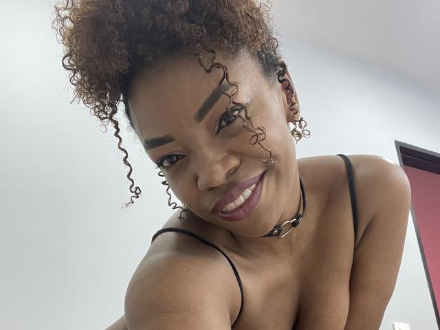 DannaBanks mit lockigem, braunem Haar, dunklen Augenbrauen, lächelnd, trägt eine schwarze Halskette und ein Top mit Trägern, zeigt ihre Brüste, hat lila Lippenstift auf und ein süßes, sexy Gesicht..