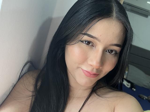 DeisyWest has long black hair, brown eyes, and a slight smile. Her tits are partially visible, showing a fair skin tone with a subtle blush on her cheeks.