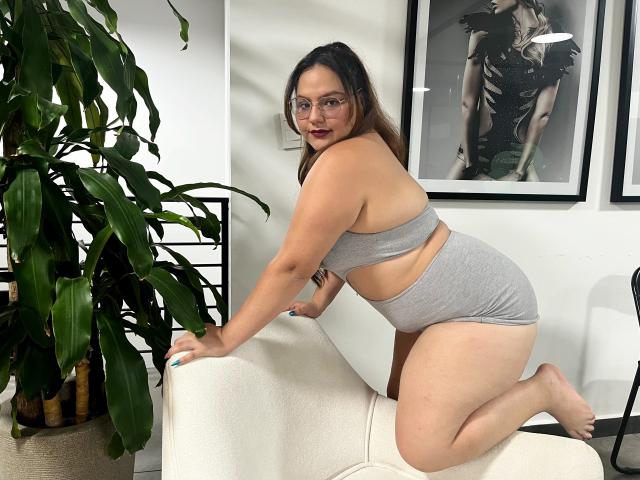 DanielleLopez, curvy, with large ass, wearing gray crop top and panties, kneeling on white chair, glasses, brown hair, looking over shoulder, smiling, indoors, plant on left.