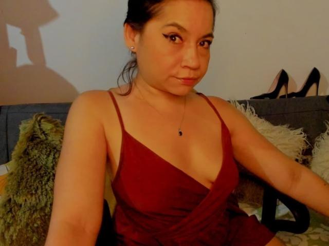 MsJustineBeaux with dark hair in a red dress showing her small tits and a subtle cleavage, sitting on a couch with black high heels in the background.