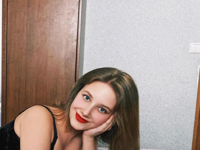 JuliaLuxx-hot with red lipstick, smiling, resting her cheek on her hand, brown hair, blue eyes, black strap dress, wooden door, grey wall, looking cute and sexy.