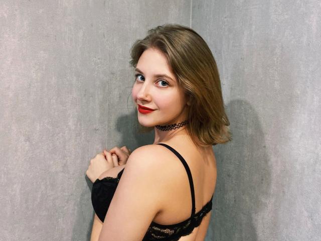 JuliaLuxx-hot with red lipstick, black lace bra, and choker, smiling over shoulder, blonde hair, pale skin, small tits, hands near chest, gray wall in background.