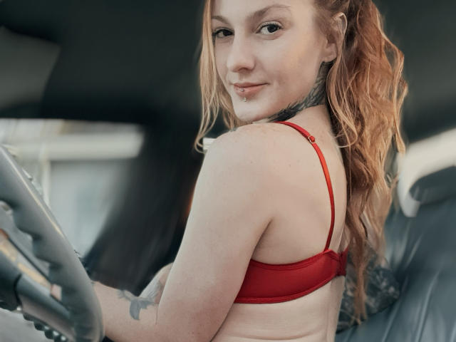 ScorpionLadyLuck with wavy red hair, red bra, tattoo on right arm, smiling at camera, holding steering wheel, subtle nose piercing, soft light, inside car, casual look.