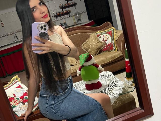 LuanaJoys with long brown hair, small boobs, wearing a white top and blue skirt, sitting, taking a mirror selfie, green and red Christmas doll on white lace table.