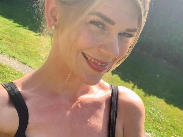 Tara-Secret-hot, blonde, smiling, winking, wearing black strap top, showing cleavage, small earrings, sunlit, grassy background, light skin, cute, attractive, casual, outdoors, selfie.