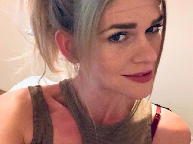 Tara-Secret-hot with blonde hair in a ponytail, wearing a brown tank top, showing her tits and red bra straps, smiling with pink lipstick and subtle makeup.