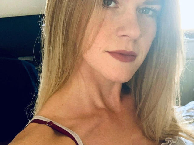 Tara-Secret-hot with long blonde hair, wearing a red strap, has a serious look, slightly parted lips, and a smooth, fair-skinned face in a car.