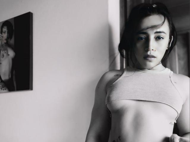 Black-and-white photo of YeiSela with short dark hair, wearing a tight, small, cropped top showing her small tits and tattoos, standing with a serious expression, ears in.