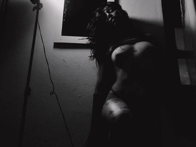 Dark, moody black-and-white photo of YeiSela with tattoos, leaning against a wall, shadowed, topless, showing tits, nipples, and a tattooed thigh, with long, messy hair.