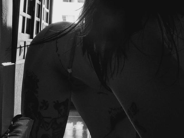 YeiSela's ass is prominent, she's leaning forward with tattoos on her thighs and a strap from a bra visible. Her face is in shadow, with long hair. Black-and-white photo.