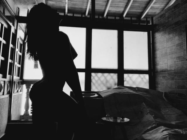 Silhouetted YeiSela with wavy hair, curvy ass, and small tits, standing in a dark room, back to the camera, near a bed with rumpled sheets. Black and white.