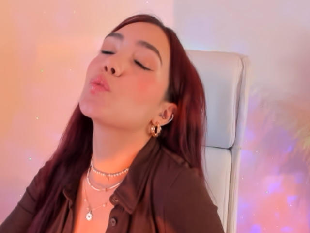 Linsay Colemann, les yeux fermés, les lèvres pincées et les cheveux roux, portant une chemise marron et un collier, assise sur une chaise blanche avec une douce lumière colorée..