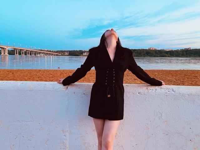 AnnSweer with her head tilted back, wearing a black dress, leaning against a white wall, hands on top, sandy beach in background, bridge on left, blue sky above.
