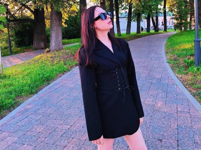 AnnSweer stands in a park wearing a black dress with lace-up detail, black blazer, and sunglasses, with her ass slightly visible, long brown hair, looking up confidently.