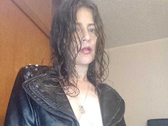 VickyHott69 with wet, dark hair, wearing a black leather jacket, showing cleavage, and a necklace, looking at the camera with slightly parted, pinkish lips.