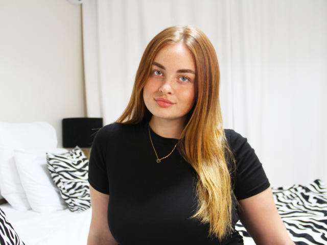 EsterMiln with long, straight, blonde hair, black t-shirt, gold necklace, cute, slightly chubby, small nose ring, standing in front of a zebra-patterned bed.