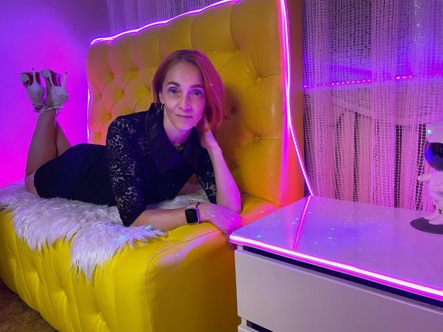 BreeTanya with pink hair, black lace dress, and high heels, lying on a yellow tufted sofa, legs up, wearing a smartwatch, under pink neon lights.