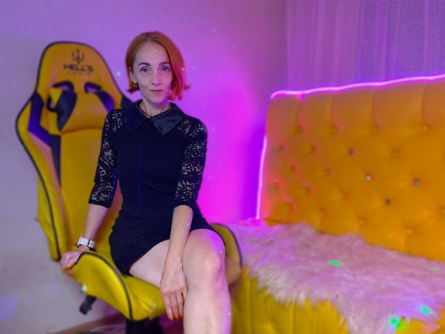 BreeTanya with short red hair, black lace dress, and watch, sitting on yellow chair, legs crossed, next to yellow tufted couch with white fur. Pink and purple lights.