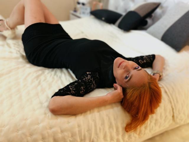BreeTanya, avec ses cheveux roux, sa robe en dentelle noire et ses talons blancs, allongée sur un lit blanc, les bras au-dessus de la tête, les jambes croisées, regardant l'appareil photo, sensuelle, détendue..