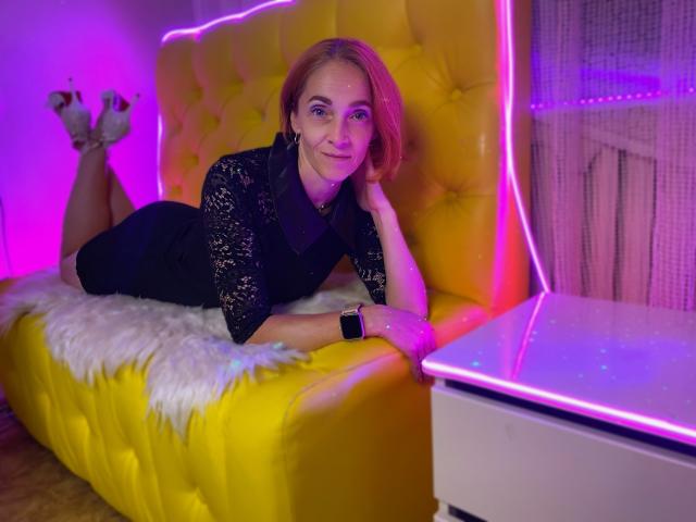 BreeTanya, com cabelo ruivo, vestindo um vestido preto de renda, deitada em um sofá amarelo com detalhes em tufos, coberta por um cobertor branco e felpudo, com luzes neon rosa e roxas, um smartwatch preto, uma mesa branca e saltos altos cinzas..