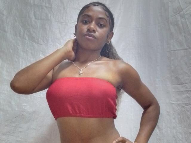 ChatBelle has dark skin, long braids, wears a red tube top, and a silver necklace with a heart pendant. She's posing with one hand on her neck, looking serious.