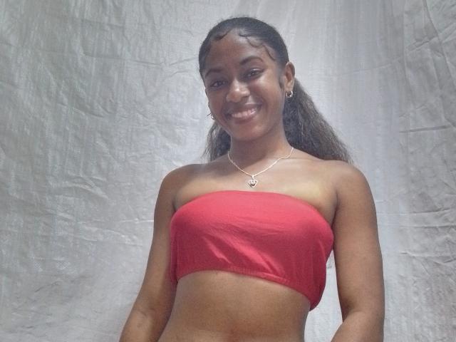 ChatBelle with medium brown skin, long brown hair, smiling, wearing a red strapless top, small silver necklace, and hoop earrings, showing her tits and flat stomach.