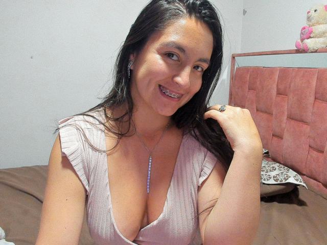 VenusSweetPalmer with long dark hair, smiling, wearing a pink ribbed top with deep cleavage, showing her large boobs, holding her hair, wearing a necklace, and a ring.
