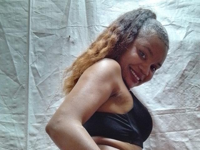 OdiJolie with dark skin, curly brown hair, black sports bra, smiling, showing her tits, standing sideways, looking over her shoulder.