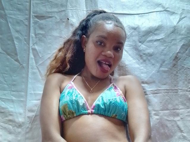 OdiJolie with brown skin, curly hair, tongue out, wearing a blue floral bikini top, small boobs, lying on a crinkled white sheet, looking at the camera.