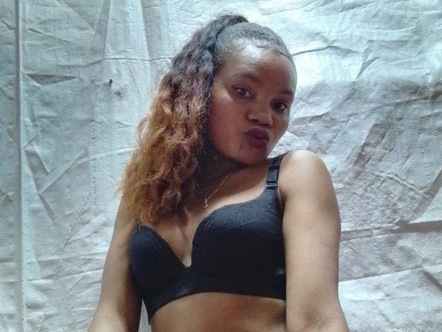 OdiJolie with dark brown skin, curly hair, and a black bra, showing her tits and a subtle pout against a crinkled white backdrop.